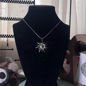 May include: A silver-toned necklace featuring a sun pendant with radiating rays. The pendant is displayed on a black velvet bust. The necklace chain is thin and delicate, complementing the detailed sunburst design.