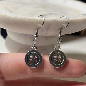 Peut inclure: Une paire de boucles d'oreilles boutons argent&eacute;es. Chaque boucle d'oreille pr&eacute;sente un petit bouton rond avec quatre trous. Les boucles d'oreilles sont suspendues &agrave; des crochets d'oreille en argent. Elles sont pr&eacute;sent&eacute;es sur un fond neutre.