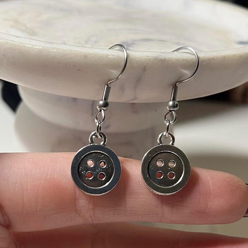 Peut inclure: Une paire de boucles d'oreilles boutons argent&eacute;es. Chaque boucle d'oreille pr&eacute;sente un petit bouton rond avec quatre trous. Les boucles d'oreilles sont suspendues &agrave; des crochets d'oreille en argent. Elles sont pr&eacute;sent&eacute;es sur un fond neutre.