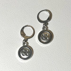 May include: A pair of silver-toned button earrings. Each earring features a circular button charm with four holes, suspended from a hinged hoop. The earrings are displayed against a white background.