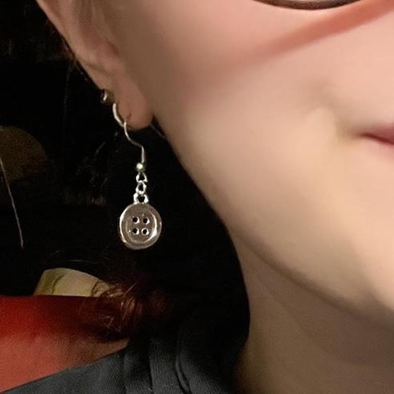 May include: A silver button earring with a hook fastening. The button has four small holes and is attached to a chain. The earring is suspended from a person's ear.