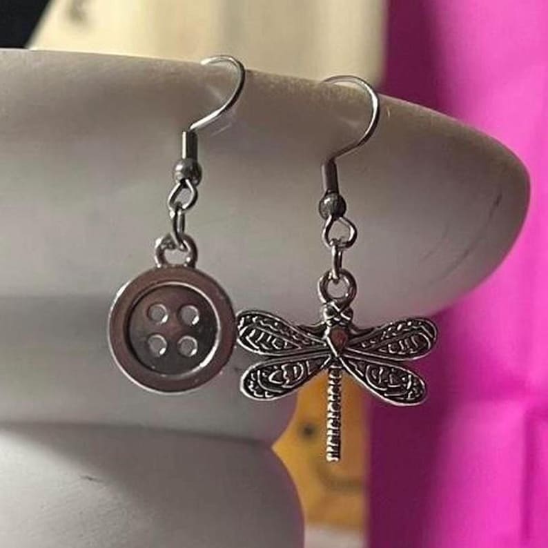May include: Silver-coloured dangle earrings. One earring has a button charm with four holes, the other a detailed dragonfly design. The earrings have a hook closure.