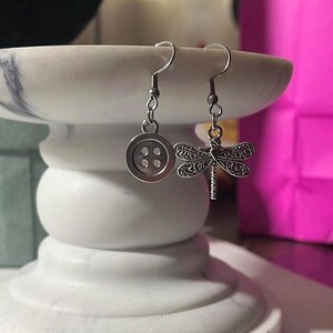May include: A pair of silver earrings. One earring has a button design with four holes, and the other features a dragonfly. The earrings hang from curved ear wires. The earrings are displayed on a white marble pedestal.