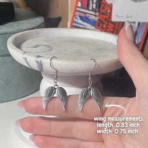 May include: Silver-colored angel wing earrings. Each wing measures 0.83 inches in length and 0.75 inches in width. The earrings are displayed on a marble-patterned surface. The image also includes text that says "wing measurements".