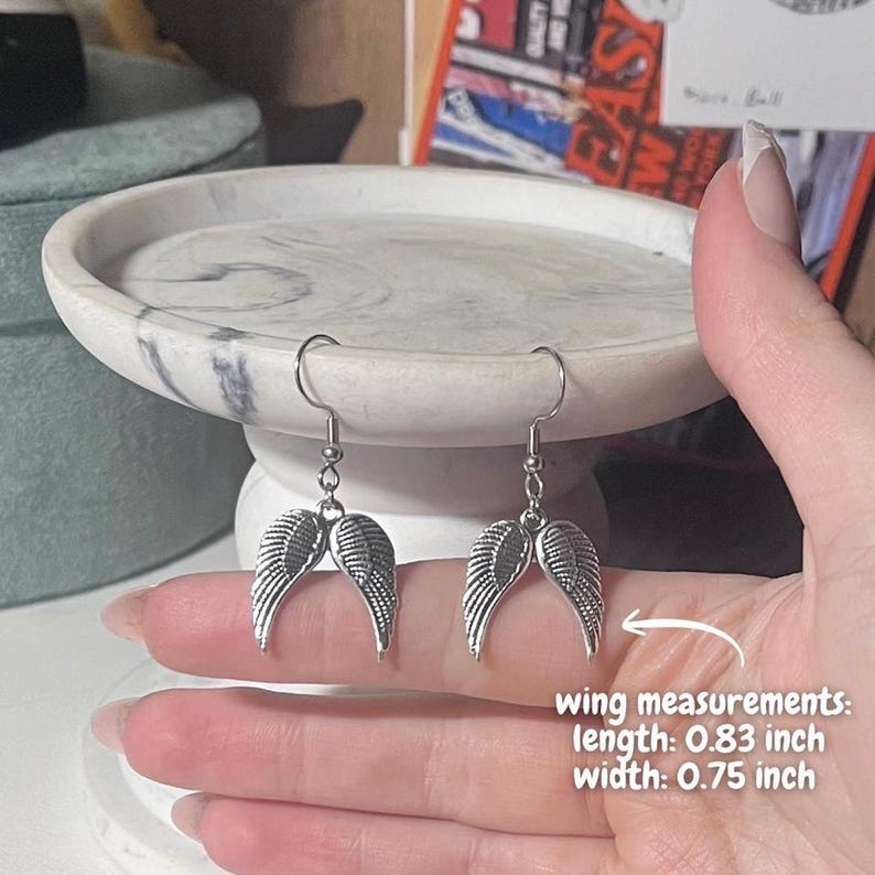 May include: Silver-colored angel wing earrings. Each wing measures 0.83 inches in length and 0.75 inches in width. The earrings are displayed on a marble-patterned surface. The image also includes text that says "wing measurements".