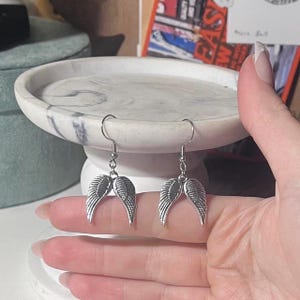 May include: A pair of silver-colored dangle earrings. Each earring features a detailed wing design. The earrings are suspended from a simple hook. The wings are displayed against a white and gray marble tray.