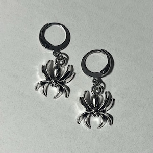 May include: A pair of silver-colored spider earrings. Each earring features a detailed spider charm suspended from a hinged hoop. The spider has a rounded body and multiple legs, creating a gothic or Halloween-themed accessory.