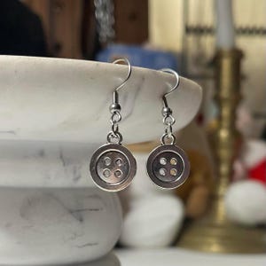 Peut inclure: Une paire de boucles d'oreilles boutons argent&eacute;es. Chaque boucle d'oreille pr&eacute;sente un charme bouton avec quatre petits trous et un crochet de poisson argent&eacute;. Les boucles d'oreilles sont pr&eacute;sent&eacute;es sur un fond flou.