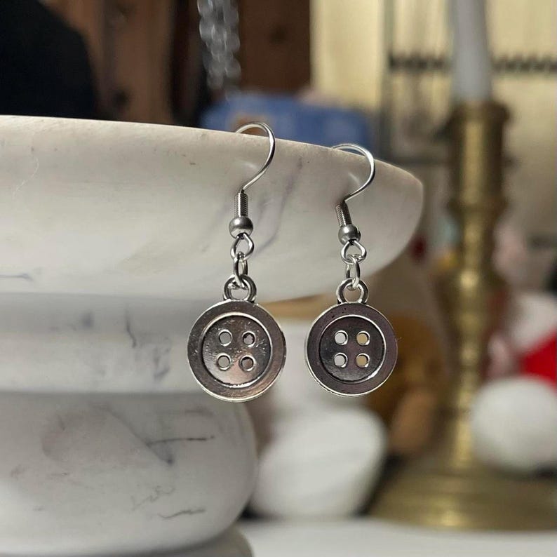 Peut inclure: Une paire de boucles d'oreilles boutons argent&eacute;es. Chaque boucle d'oreille pr&eacute;sente un charme bouton avec quatre petits trous et un crochet de poisson argent&eacute;. Les boucles d'oreilles sont pr&eacute;sent&eacute;es sur un fond flou.