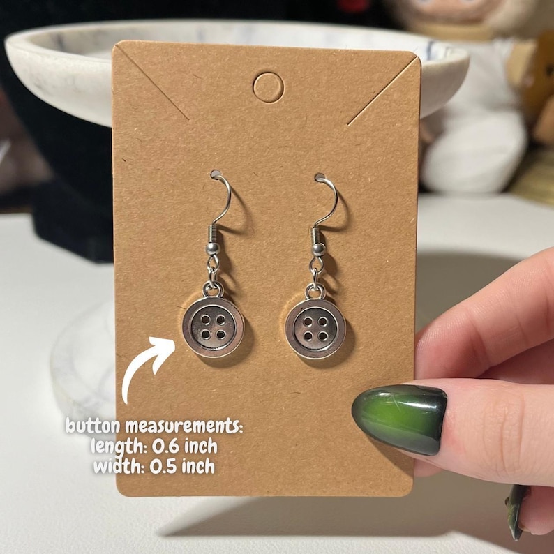 Peut inclure: Boucles d'oreilles boutons argent&eacute;es avec un design &agrave; quatre trous. Chaque bouton mesure 1,5 cm de long et 1,3 cm de large. Les boucles d'oreilles sont suspendues &agrave; un crochet simple. Elles sont pr&eacute;sent&eacute;es sur une carte brune.