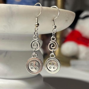 May include: Silver-toned dangle earrings. Each earring features a button charm with four holes, connected to a spiral design and a hook. The earrings are displayed against a blurred background.