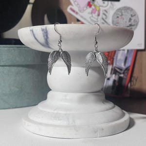 May include: A pair of silver-colored angel wing earrings. The earrings are displayed on a white marble pedestal. The wings are detailed with feather-like textures and hang from silver hooks.