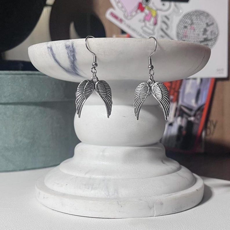 May include: A pair of silver-colored angel wing earrings. The earrings are displayed on a white marble pedestal. The wings are detailed with feather-like textures and hang from silver hooks.
