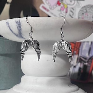 May include: A pair of silver-colored earrings featuring a wing design. Each earring has a curved wing shape with detailed feather textures, suspended from a simple hook. The earrings are displayed on a white marble surface.