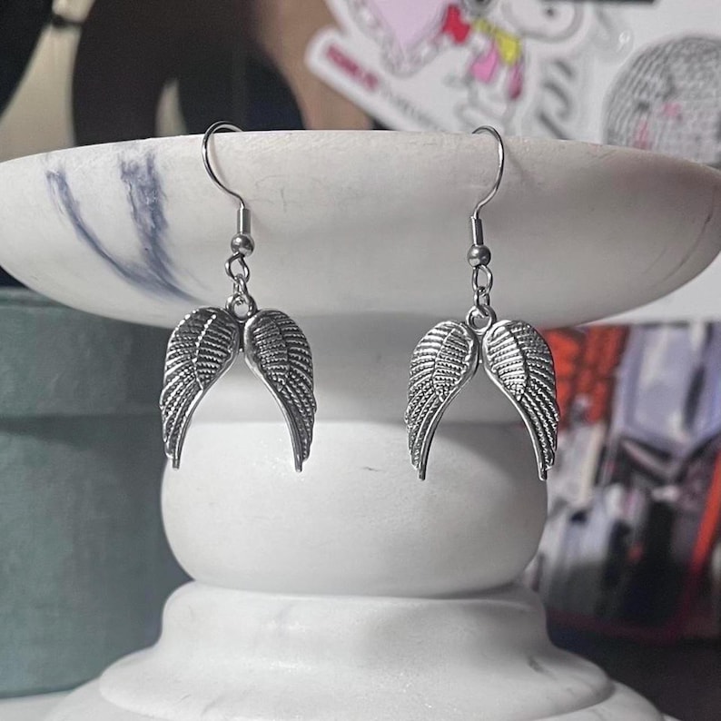 May include: A pair of silver-colored earrings featuring a wing design. Each earring has a curved wing shape with detailed feather textures, suspended from a simple hook. The earrings are displayed on a white marble surface.