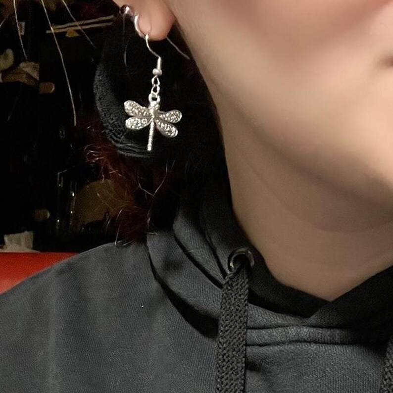May include: A silver dragonfly earring is displayed, featuring detailed wings. The earring hangs from a small hoop. The dragonfly design is the main focus, with intricate detailing. The earring is worn on the ear.