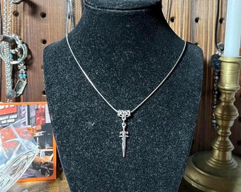 May include: A silver chain necklace with a silver dagger pendant. The necklace is displayed on a black mannequin bust.