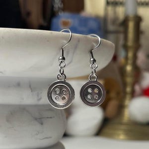 May include: A pair of silver button earrings. Each earring features a small, round button charm with four holes, suspended from a simple hook. The earrings are displayed against a blurred background.