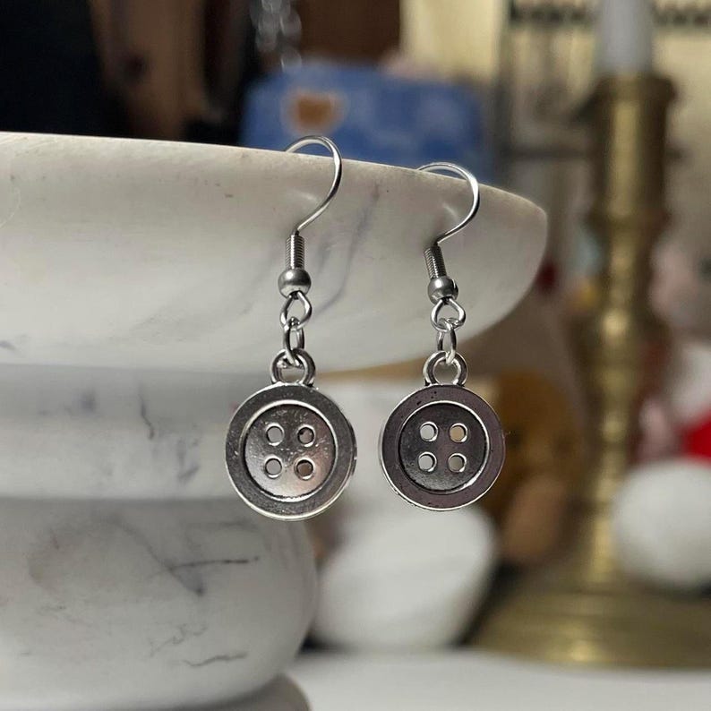 Peut inclure: Une paire de boucles d'oreilles boutons argent&eacute;es. Chaque boucle d'oreille pr&eacute;sente un petit bouton rond avec quatre trous, suspendu &agrave; un crochet simple. Les boucles d'oreilles sont pr&eacute;sent&eacute;es sur un fond flou.