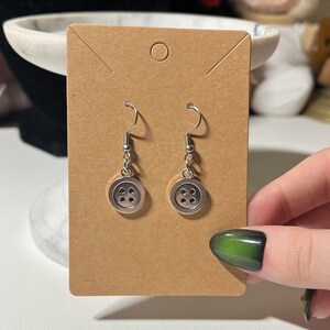 Peut inclure: Une paire de boucles d'oreilles bouton avec des crochets et des breloques en forme de bouton argent&eacute;s. Chaque breloque bouton comporte quatre petits trous. Les boucles d'oreilles sont pr&eacute;sent&eacute;es sur une carte marron.