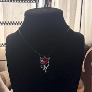 May include: A necklace featuring a silver-toned metal pendant shaped like a heart with small devil horns and a tail, filled with a glossy red enamel center. The pendant hangs from a simple black cord, displayed on a black mannequin bust.