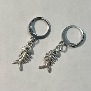 May include: A pair of silver-colored hoop earrings. Each earring features a fish skeleton charm dangling from a hinged hoop. The fish skeleton charms have detailed rib structures and a small hole for the eye.