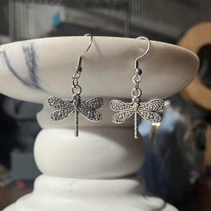 May include: A pair of silver-colored dragonfly earrings. Each earring features a detailed dragonfly charm with textured wings and a delicate, dangling design. The earrings are suspended from simple, curved ear wires.