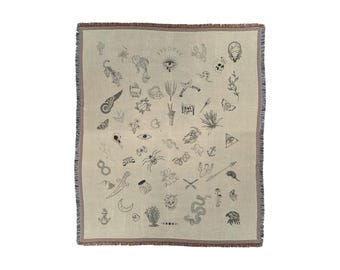 May include: A beige woven throw blanket with a brown fringe border. The blanket features a pattern of various black and white tattoo-style illustrations, including a skull, an eye, a snake, a bird, a heart, a flower, and a crescent moon.