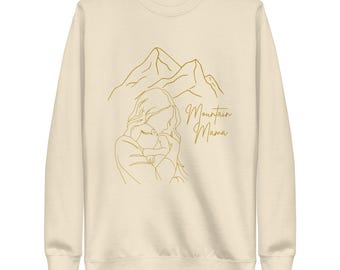 May include: A cream colored sweatshirt with a gold outline of a mountain range and a mother holding a child. The text "Mountain Mama" is written in gold script.