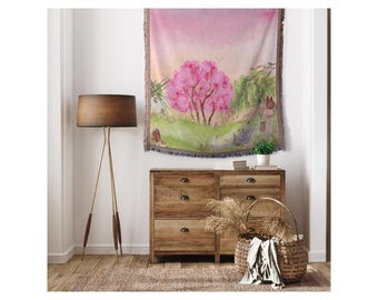 May include: A woven tapestry wall hanging with a watercolor painting of two pink flowering trees in a green field. The tapestry has a fringe border and is hanging on a white wall above a wooden dresser.