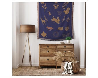 May include: A navy blue throw blanket with a gold pattern of animals and plants. The blanket is hanging on a wall above a wooden dresser with drawers. A wicker basket with a white cloth inside is sitting on the floor in front of the dresser.