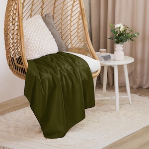 May include: A dark green throw blanket draped over a wicker chair with a white cushion. The chair is in a room with a white side table and a vase of white flowers.