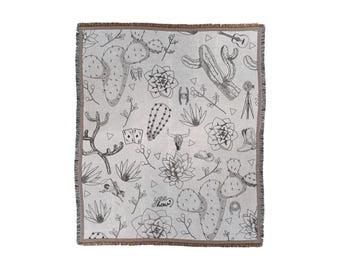 May include: A woven throw blanket with a white background and black line drawings of cacti, succulents, cowboy boots, a guitar, a skull, dice, and the words "yee haw".