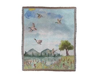 May include: A woven throw blanket with a watercolor design of ducks flying over a lake with green trees and reeds. The sky is blue with white clouds and a yellow sun.