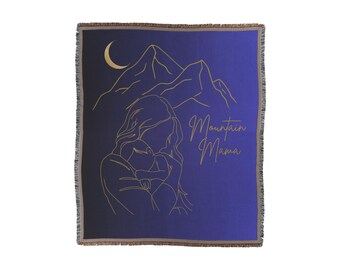 May include: A navy blue throw blanket with a gold outline of a woman holding a child, mountains, and a crescent moon. The text "Mountain Mama" is written in gold.