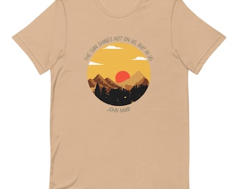 May include: A tan t-shirt with a graphic of a sunset over mountains in a circle. The text "The sun shines not on us but in us" is around the circle. The text "John Muir" is below the circle.