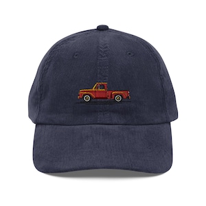 May include: Navy blue corduroy baseball cap featuring an embroidered red vintage pickup truck design. The truck has yellow headlights and wheels. The cap has a curved brim and a button on top.