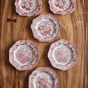 May include: Six pink and white floral patterned plates arranged on a wooden surface. The plates have a scalloped edge and a detailed floral design in shades of pink and white. The plates are arranged in a circular pattern.