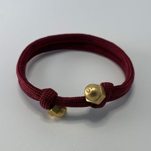 Op de afbeelding: Een maroon paracord armband met twee gouden metalen accenten. De accenten zijn klein, rond en hebben een reliëfletter 'S' erop.