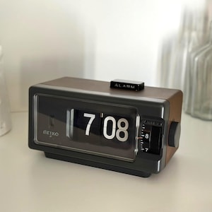 May include: A rectangular Seiko flip clock with a dark wood-grain finish and a black front panel. The display shows the time as 7:08 in white numerals. A black alarm switch is located on the top, and a circular dial is on the right side.