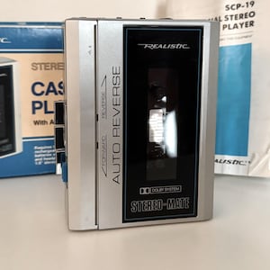 May include: A silver Realistic Stereo-Mate cassette player with auto reverse, featuring a black central panel with white text. The device is displayed alongside its original blue and white cardboard packaging box and a white instruction manual.