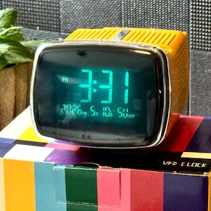 May include: A vintage yellow and black VFD clock displaying 3:31 AM in green digits. The clock sits atop a colorful box with vertical stripes of red, blue, and yellow. A potted plant is in the background.