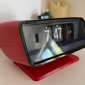 May include: A red Seiko flip clock with a black rectangular face and a clear protective cover. The digital display shows the time as 7:47 with white numbers on black tiles. A small black alarm switch is located on the top of the red plastic casing.