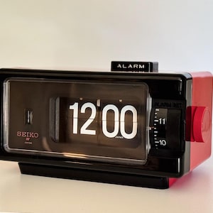 Puede incluir: Un reloj de solapa Seiko rectangular con panel frontal negro y carcasa roja. La pantalla muestra la hora 12:00 en números blancos. Un dial negro de ajuste de alarma está en el lado derecho y un botón negro ALARM está en la parte superior.