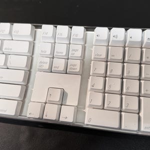 May include: A white computer keyboard with a numeric keypad and function keys. The keyboard has white keycaps with black lettering. The keys include numbers, letters, and symbols. The keyboard is on a dark surface.