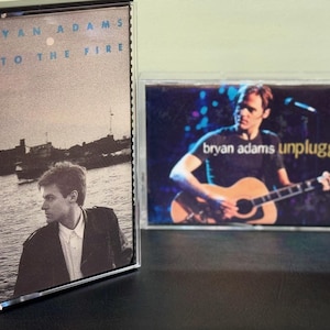 Könnte beinhalten: Zwei Musikkassetten von Bryan Adams. Eine zeigt das Album "Into the Fire" mit einem Schwarz-Weiß-Foto von Adams. Die andere ist "Unplugged" mit Adams, der Gitarre spielt.