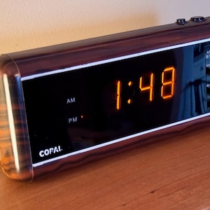 May include: A vintage Copal digital alarm clock with a wood-grain finish. The time displayed is 1:48 in bright orange digits. The clock has a black face with white trim and control buttons on the top and side. The clock is sitting on a wooden surface.