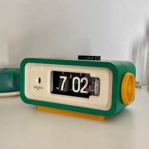 May include: A rectangular Seiko flip clock with a green outer casing, a cream-coloured face, and a yellow base and side dial. The digital display shows the time as 7:02 PM in white numerals against a black background. A black alarm switch is on top.