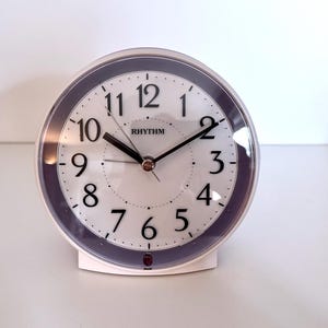 May include: A white and purple analog alarm clock with a clear plastic cover. The clock face has black numerals and hands, with the brand name "RHYTHM" printed at the top. The clock is on a white surface.