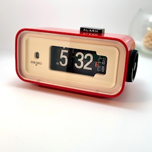 Puede incluir: Un reloj de solapa Seiko rectangular con una carcasa exterior roja brillante y una cara de color crema. La pantalla digital muestra la hora 5:32 PM con paneles abatibles negros y números blancos. Un interruptor de alarma negro está en la parte superior.